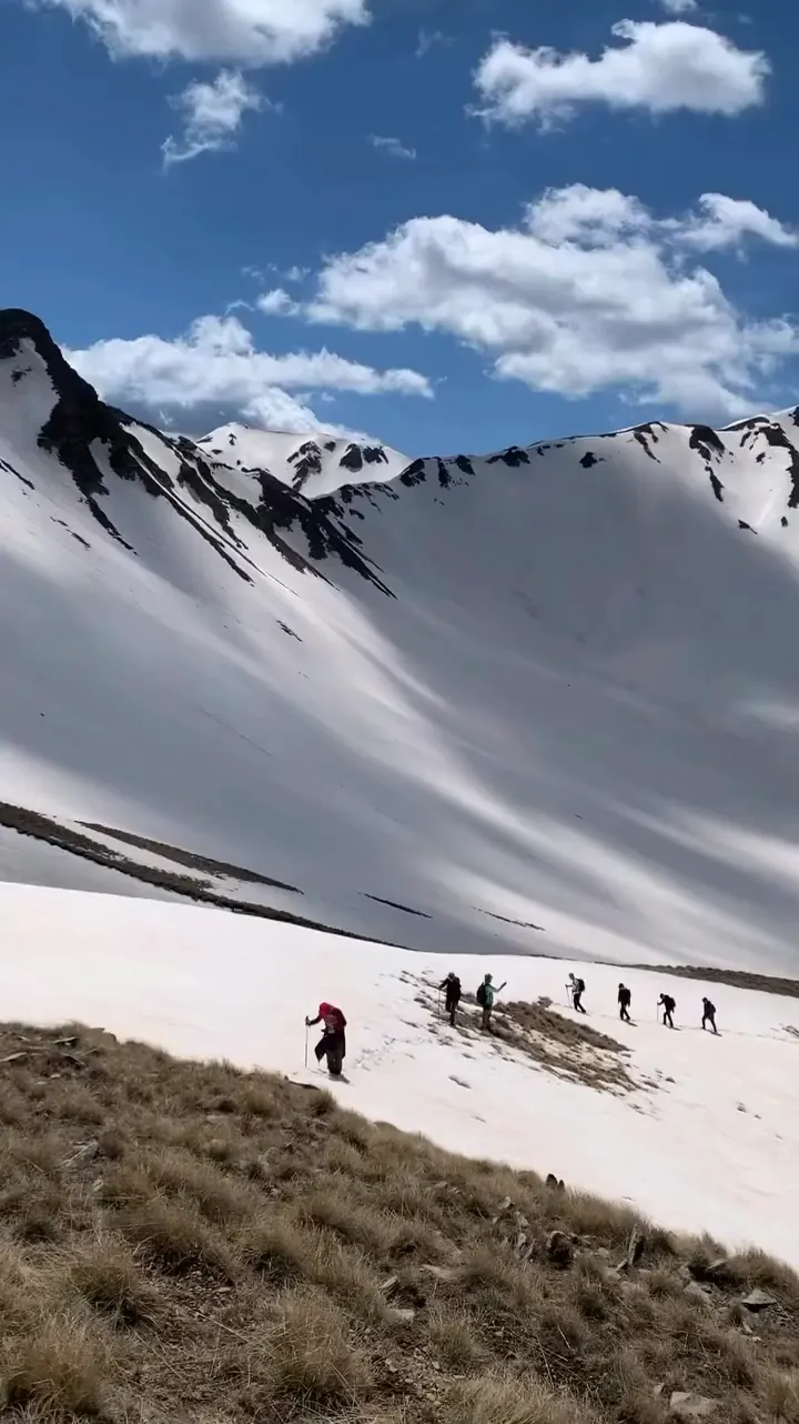 Hikers crossing a broad snowy mountain slope beneath a blue sky